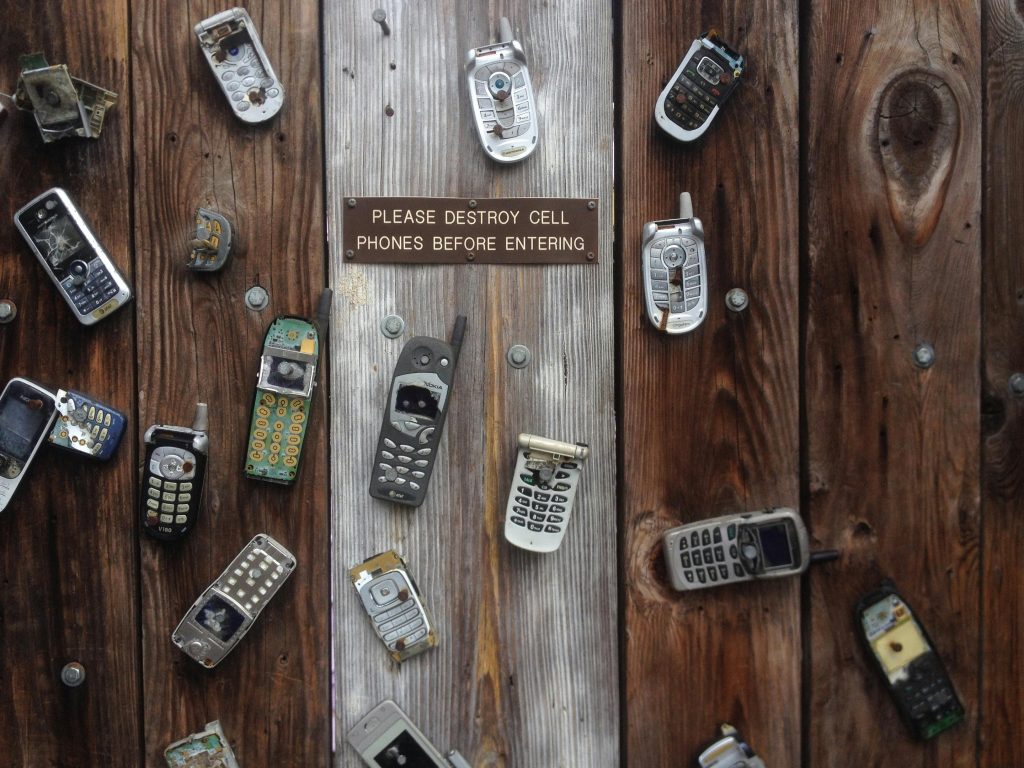 A tabletop covered in broken cell phones and a sign saying ‘Please destroy cell phones before entering,’ urging you to declutter your digital life.